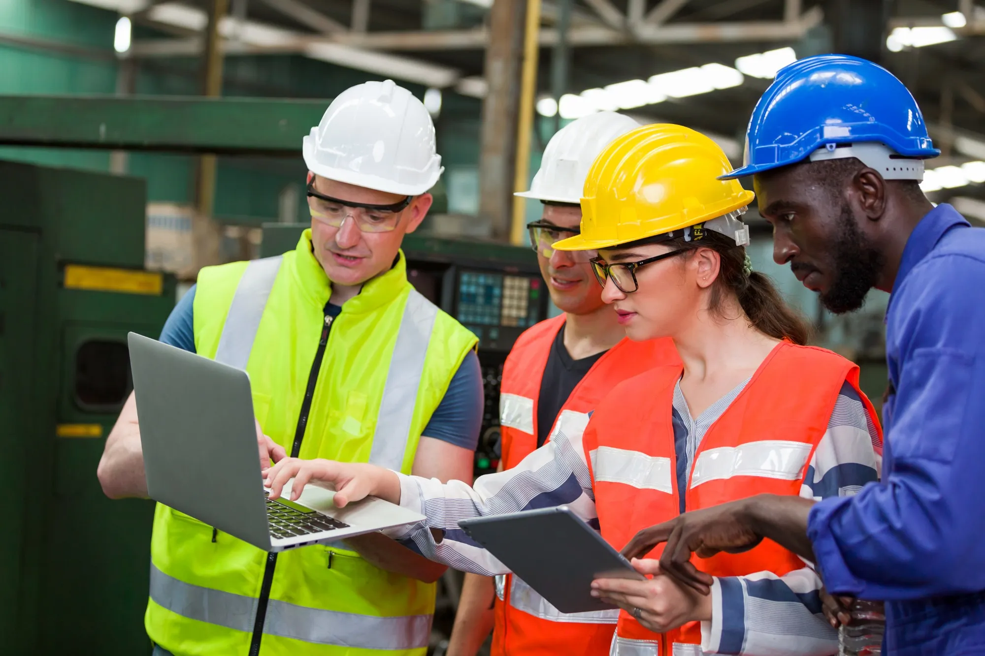 Four production workers in hard hats and safety vests review data together on a laptop and a tablet on the factory floor